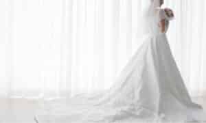 immagine di una sposa con indosso il suo abito davanti alla tenda bianca di una finestra
